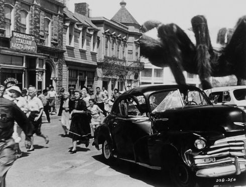 Black-and-white film still of a giant spider looming over a town street while people run past a parked car